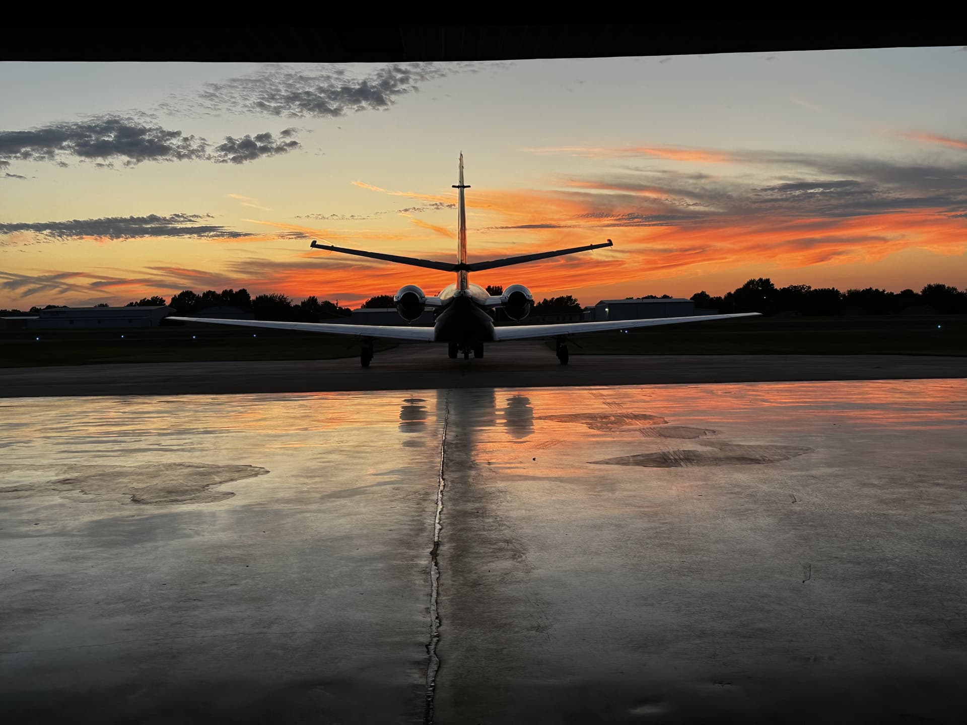 A business jet silhouetted head-on against a fiery sunset, reflected on the wet ramp